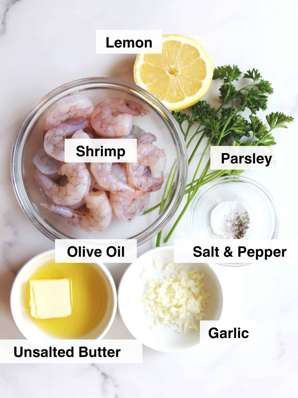 Top-down view of labeled ingredients for garlic shrimp: raw shrimp, half a lemon, parsley, minced garlic, olive oil, unsalted butter, and a small bowl of salt & pepper on a white marble countertop.