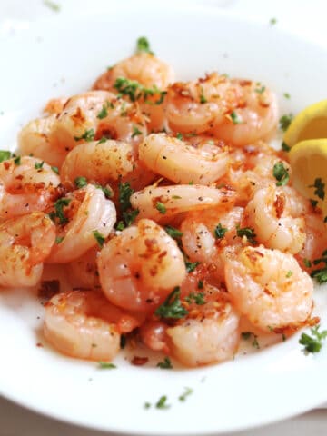 A plate of cooked shrimp garnished with chopped parsley and crispy garlic bits, served with two lemon wedges on the side.