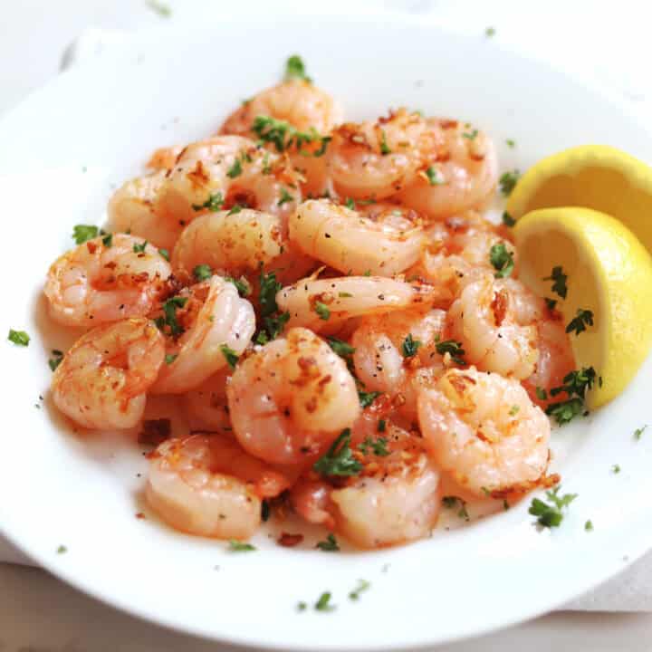 A plate of cooked shrimp garnished with chopped parsley and crispy garlic bits, served with two lemon wedges on the side.