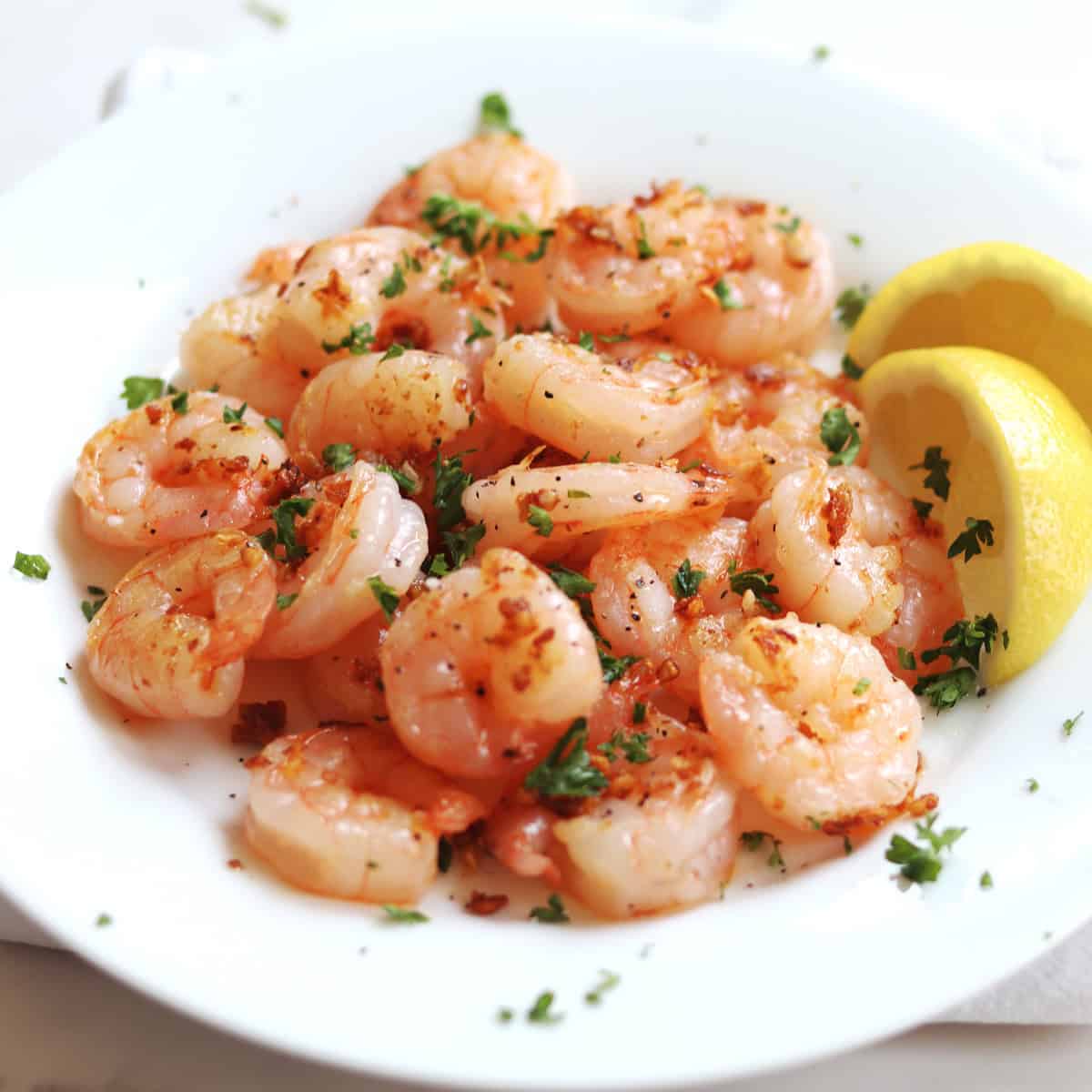 A plate of cooked shrimp garnished with chopped parsley and crispy garlic bits, served with two lemon wedges on the side.