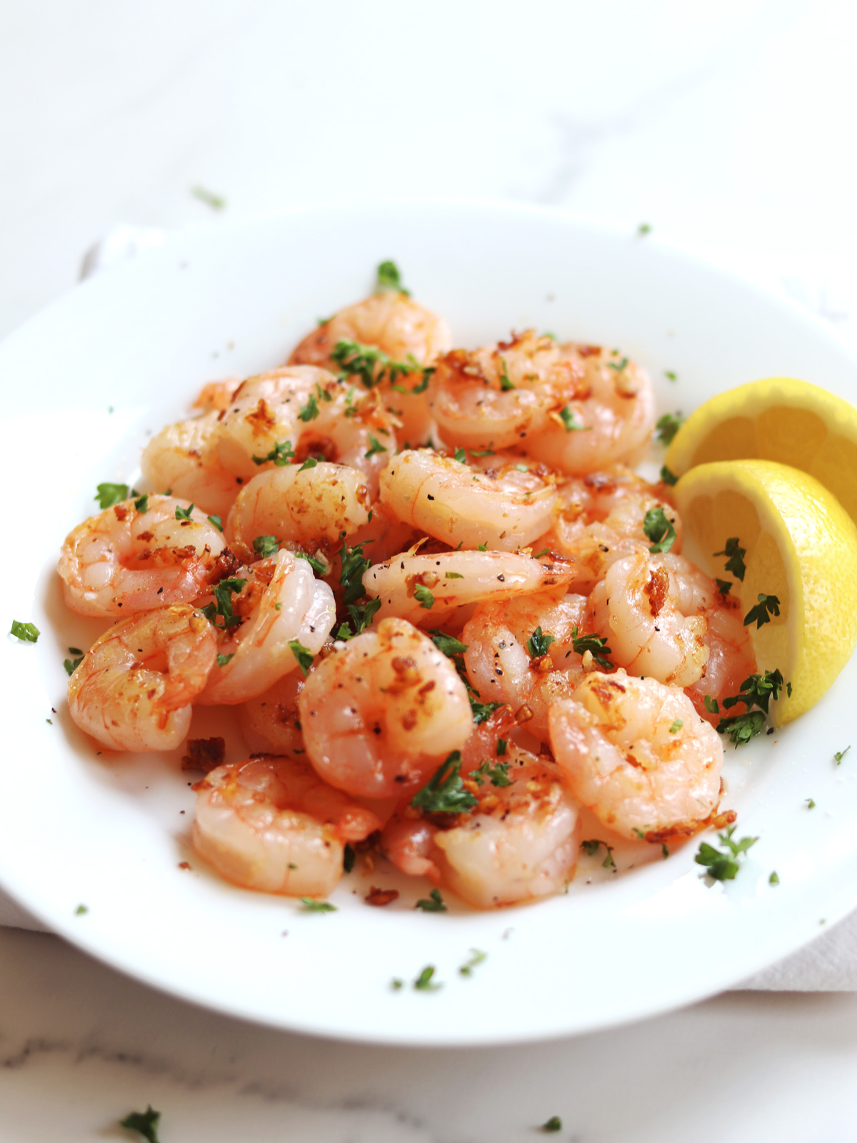 A white plate of cooked shrimp garnished with chopped parsley and crispy garlic bits, served with two lemon wedges on the side.