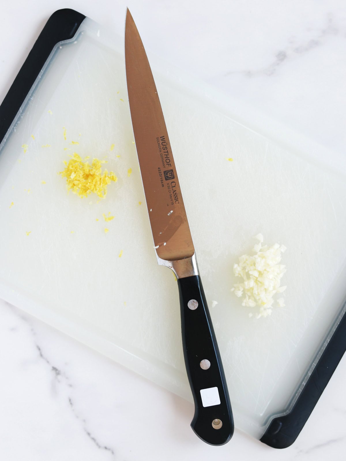 A chefs knife rests on a white cutting board with small piles of minced garlic on the right and lemon zest on the left.