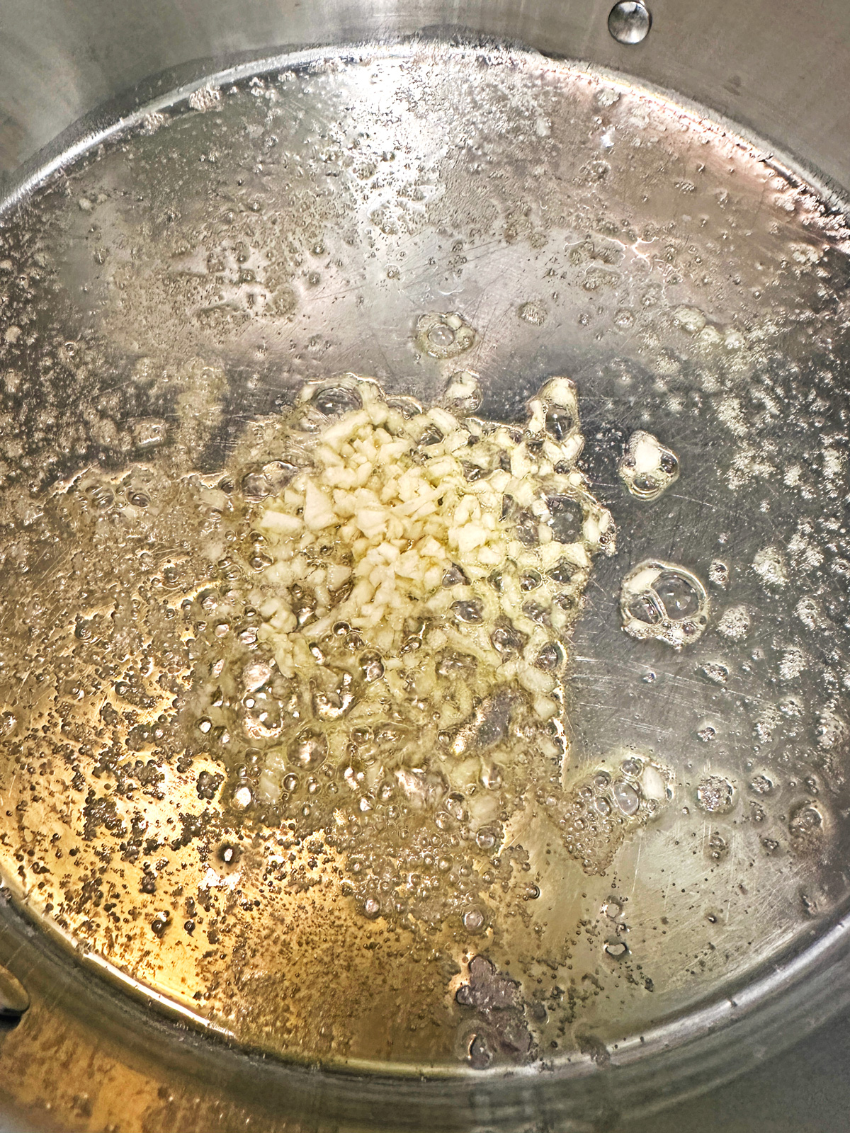 Chopped garlic sizzling in hot oil and butter in a stainless steel skillet, beginning to brown and release aroma, with bubbles visible around the garlic pieces.