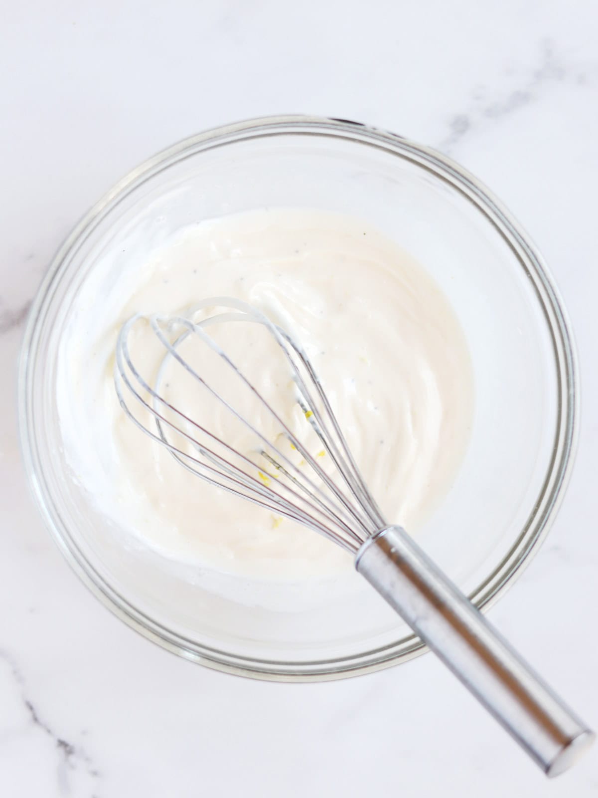 A metal whisk rests in a glass bowl filled with garlic aioli sauce on a white marble countertop.