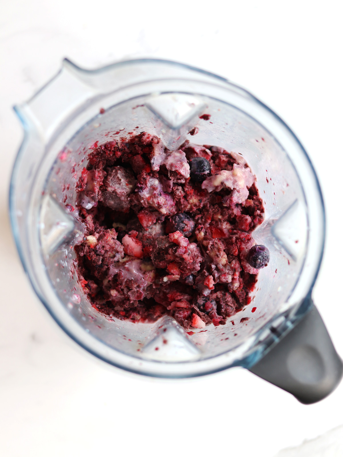 A top-down view of a blender containing a mixture of chopped frozen berries, acai and banana pieces, partially blended, with visible chunks and a creamy texture.