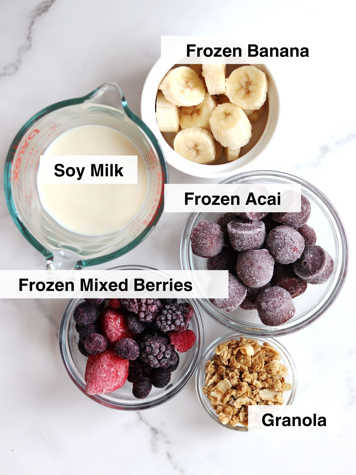 Five clear bowls and cups on a white surface contain soy milk, frozen banana slices, frozen acai puree, frozen mixed berries, and granola, each labeled with text.