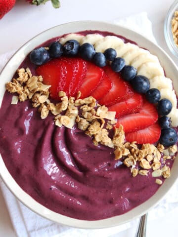 An acai bowl topped with sliced bananas, strawberries, blueberries, and granola. A white napkin and granola bowl are nearby.