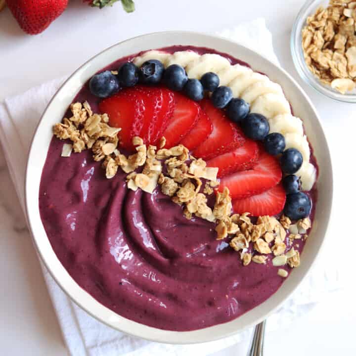 An acai bowl topped with sliced bananas, strawberries, blueberries, and granola. A white napkin and granola bowl are nearby.
