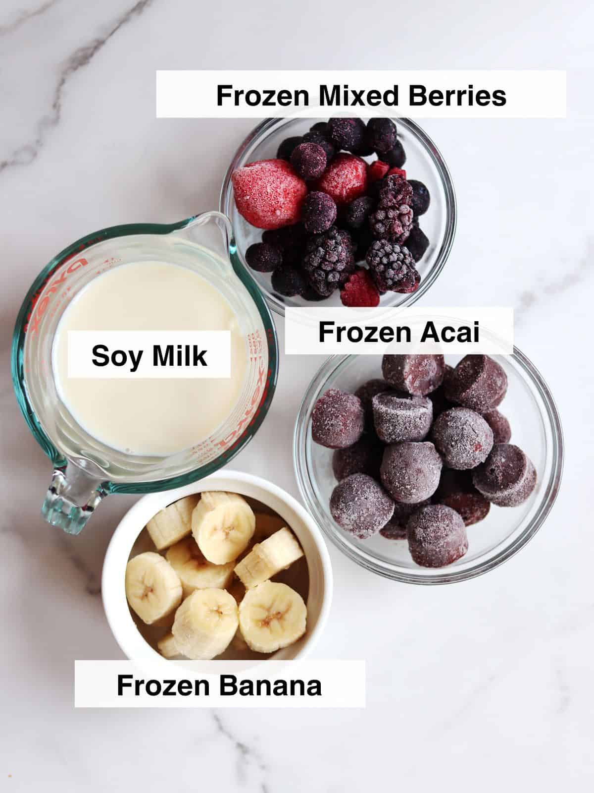 Four bowls on a marble countertop filled with soy milk, frozen mixed berries, frozen acai, and sliced frozen bananas, prepared for making a smoothie.