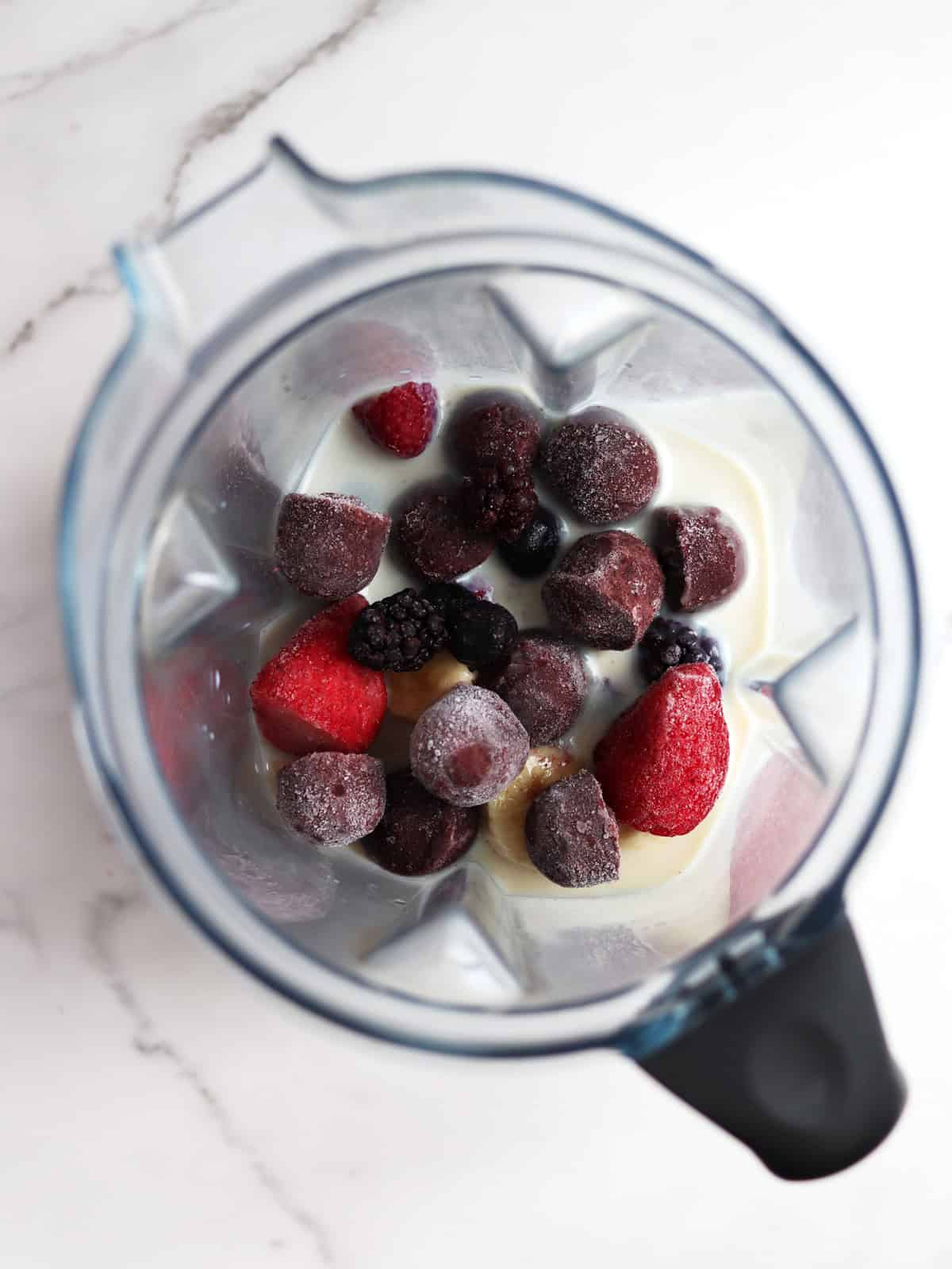 A blender with frozen acai, mixed berries and banana slices in soy milk, viewed from above on a white marble countertop.