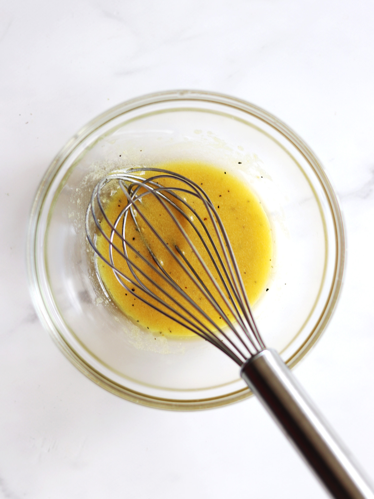 A metal whisk rests in a glass bowl containing a fresh lemon vinaigrette on a white countertop.