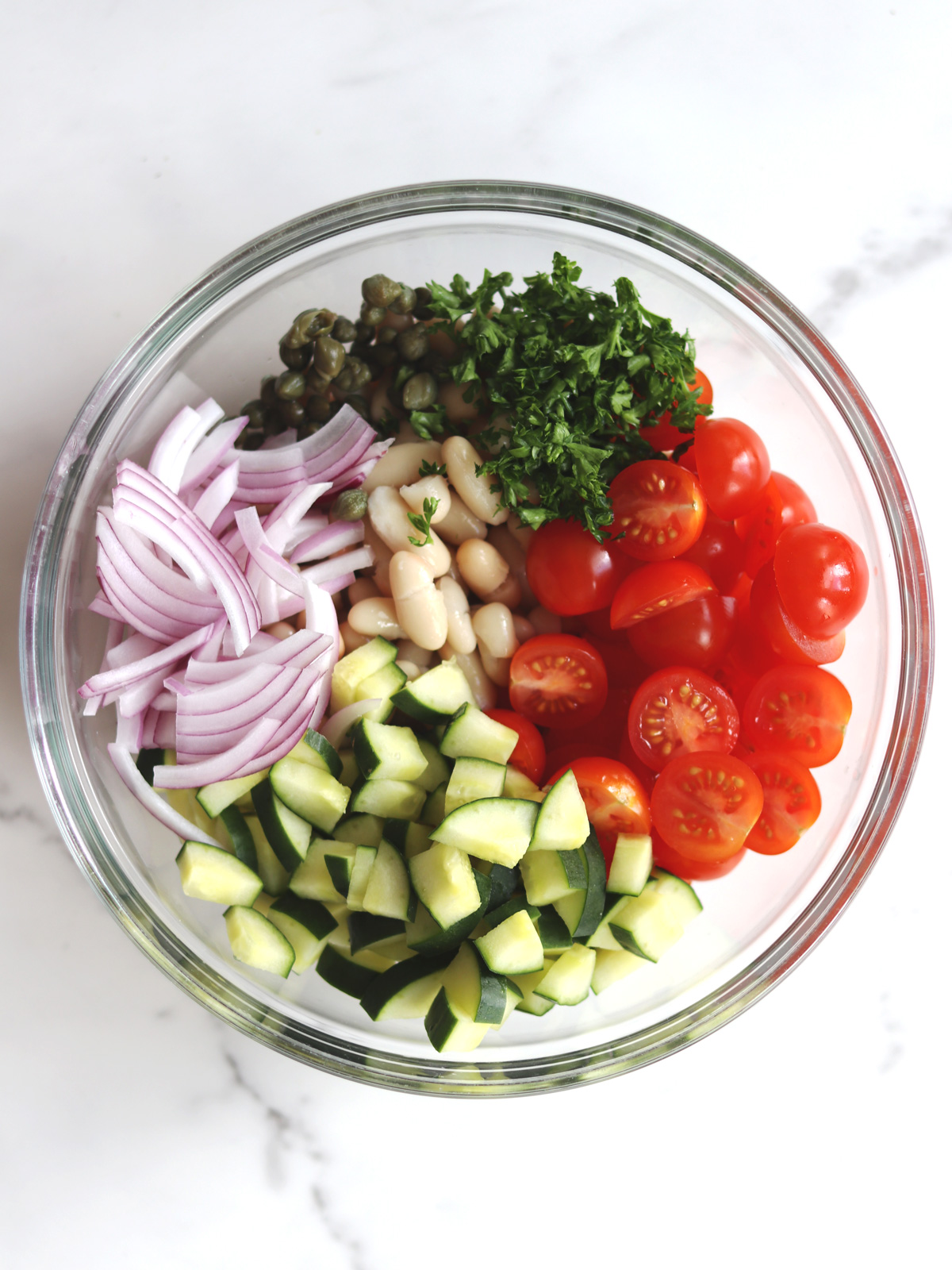 A glass bowl containing sliced red onions, capers, chopped parsley, grape tomatoes, cannellini beans, and diced cucumber, arranged in separate sections on a white marble countertop.
