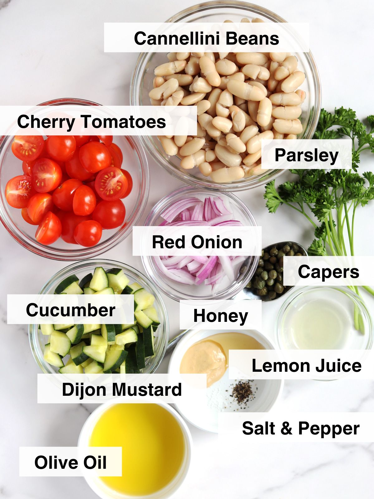 Top-down view of labeled ingredients in bowls for a white bean salad: cannellini beans, cherry tomatoes, cucumber, red onion, parsley, capers, honey, lemon juice, Dijon mustard, olive oil, and salt & pepper.