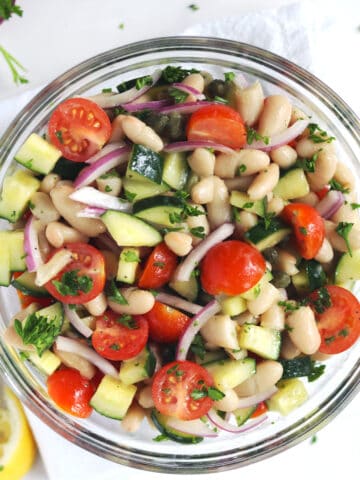 A glass bowl filled with a white bean salad made with chopped cucumber, halved cherry tomatoes, sliced red onion, capers, and fresh parsley, placed on a white napkin with squeezed lemon slices nearby.
