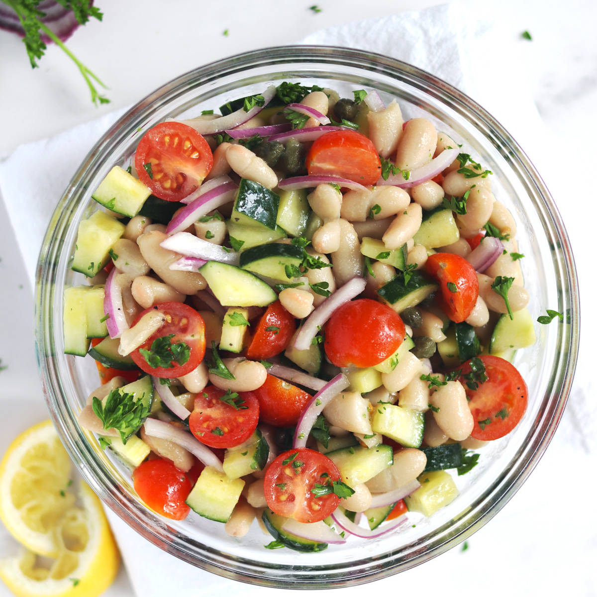 A glass bowl filled with a white bean salad made with chopped cucumber, halved cherry tomatoes, sliced red onion, capers, and fresh parsley, placed on a white napkin with squeezed lemon slices nearby.