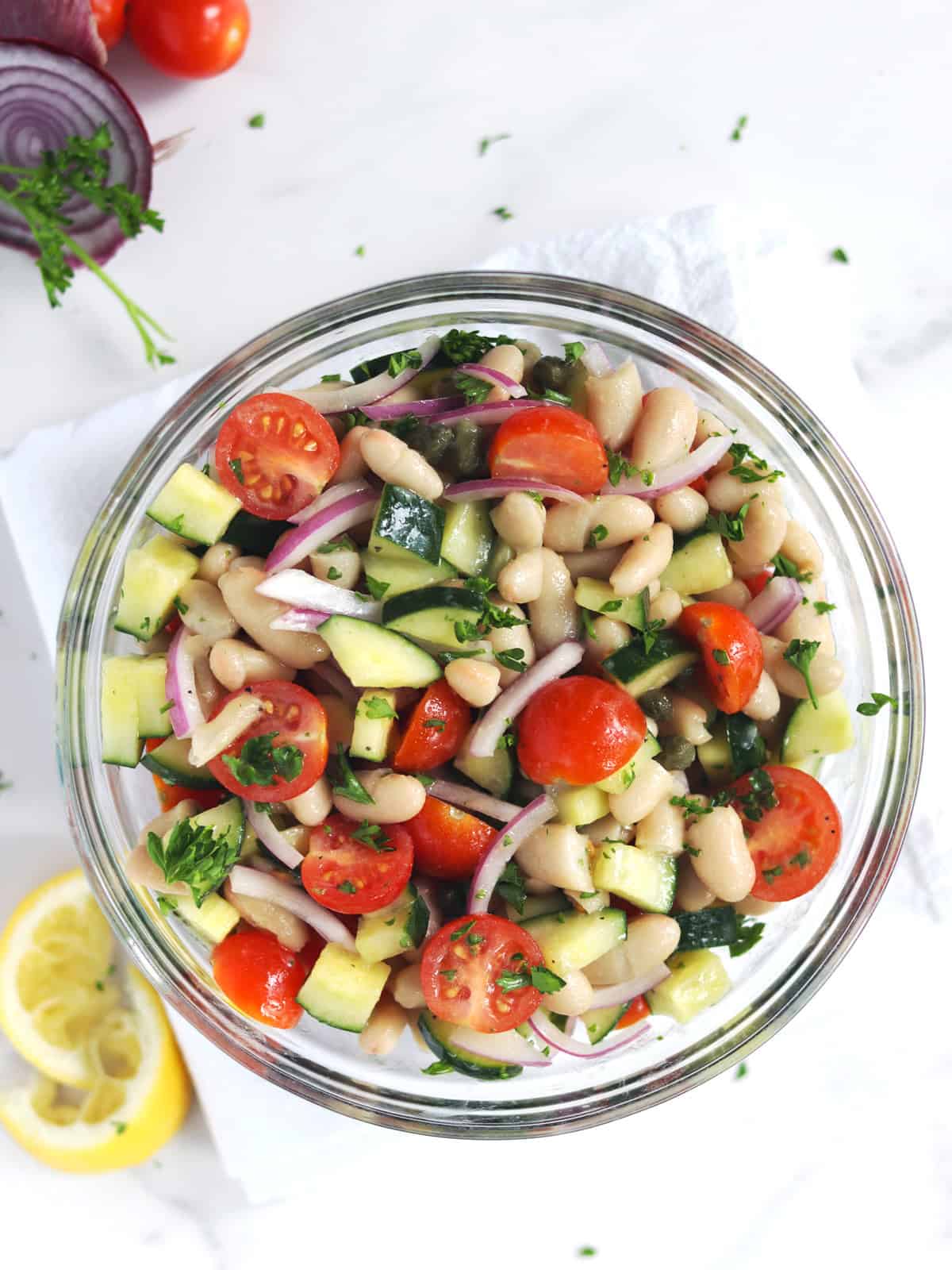 A glass bowl filled with a fresh white bean salad including cherry tomatoes, cucumber, capers, red onion, and chopped parsley, placed on a white napkin with lemon slices and cherry tomatoes nearby.