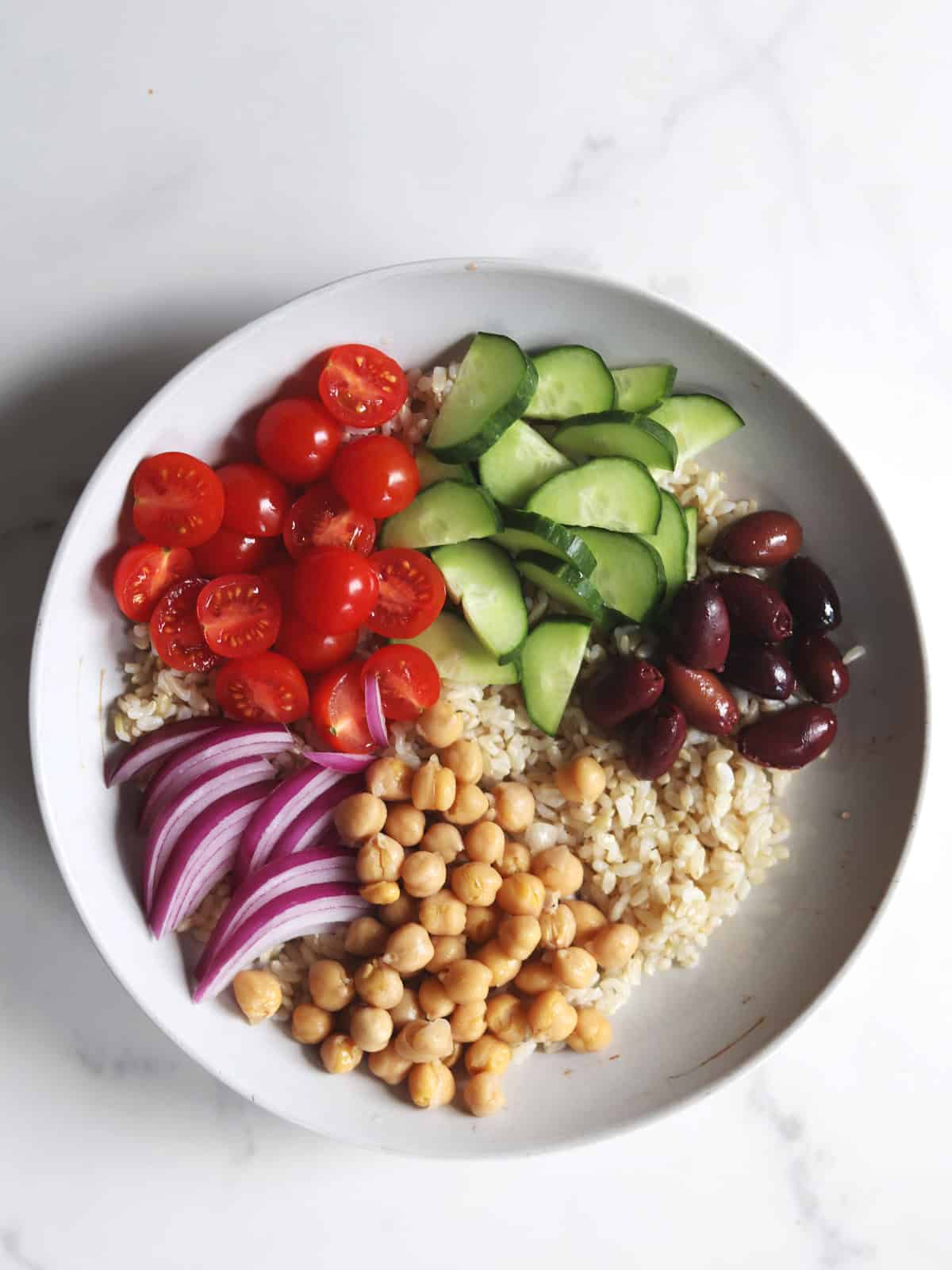 A gray ceramic bowl with brown rice, sliced cucumbers, cherry tomatoes, Kalamata olives, red onion slices, and chickpeas, arranged in separate sections.
