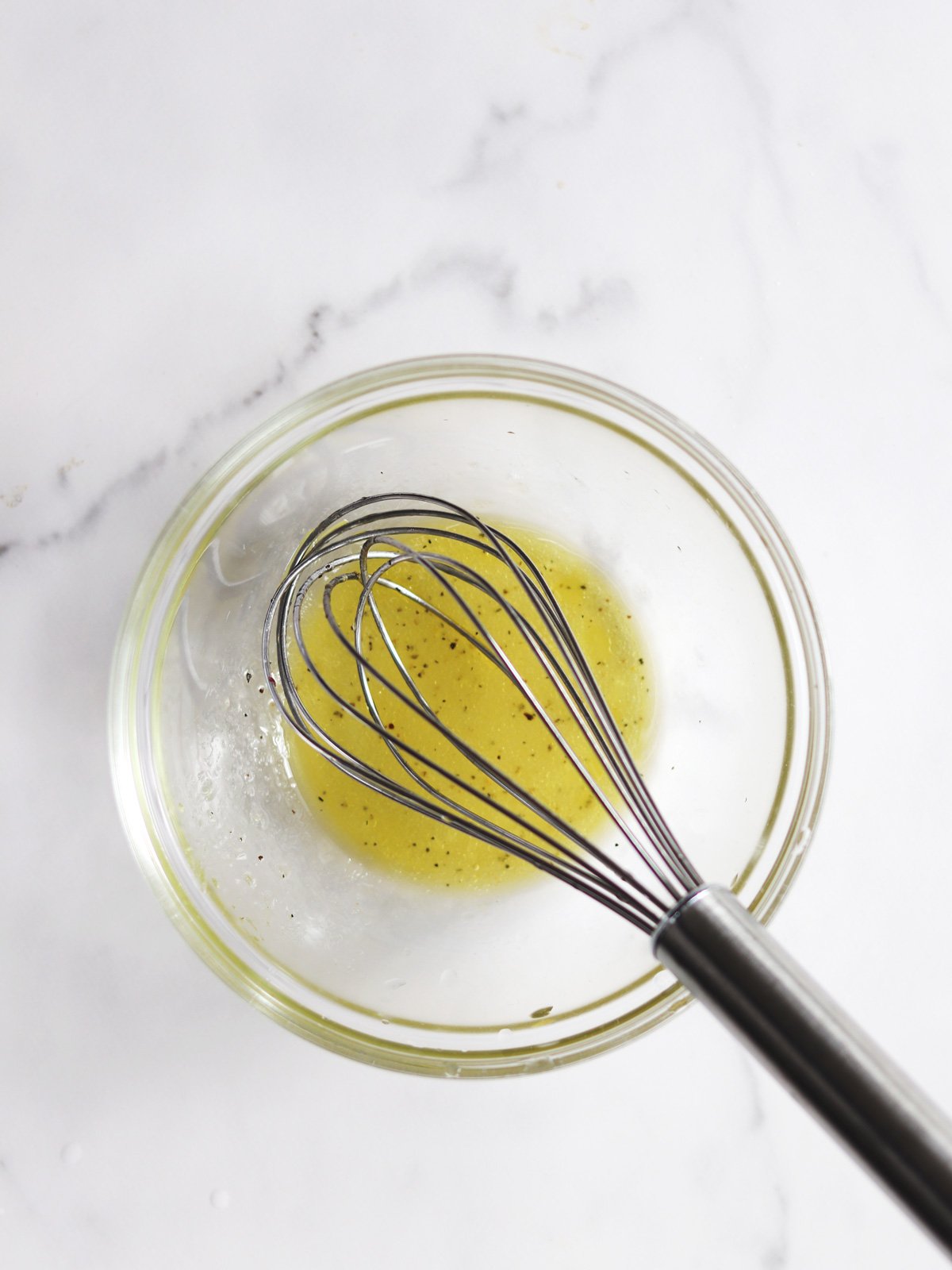 A glass bowl with lemon vinaigrette and black pepper specks being whisked by a metal whisk, on a white marble countertop.