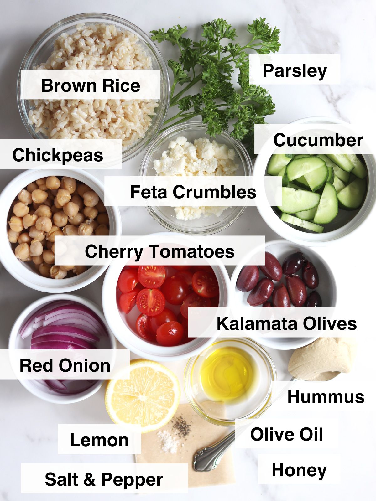Bowls of brown rice, chickpeas, feta, cucumber, cherry tomatoes, red onion, kalamata olives, hummus, lemon, olive oil, honey, salt, pepper, and parsley on a white surface, each labeled.