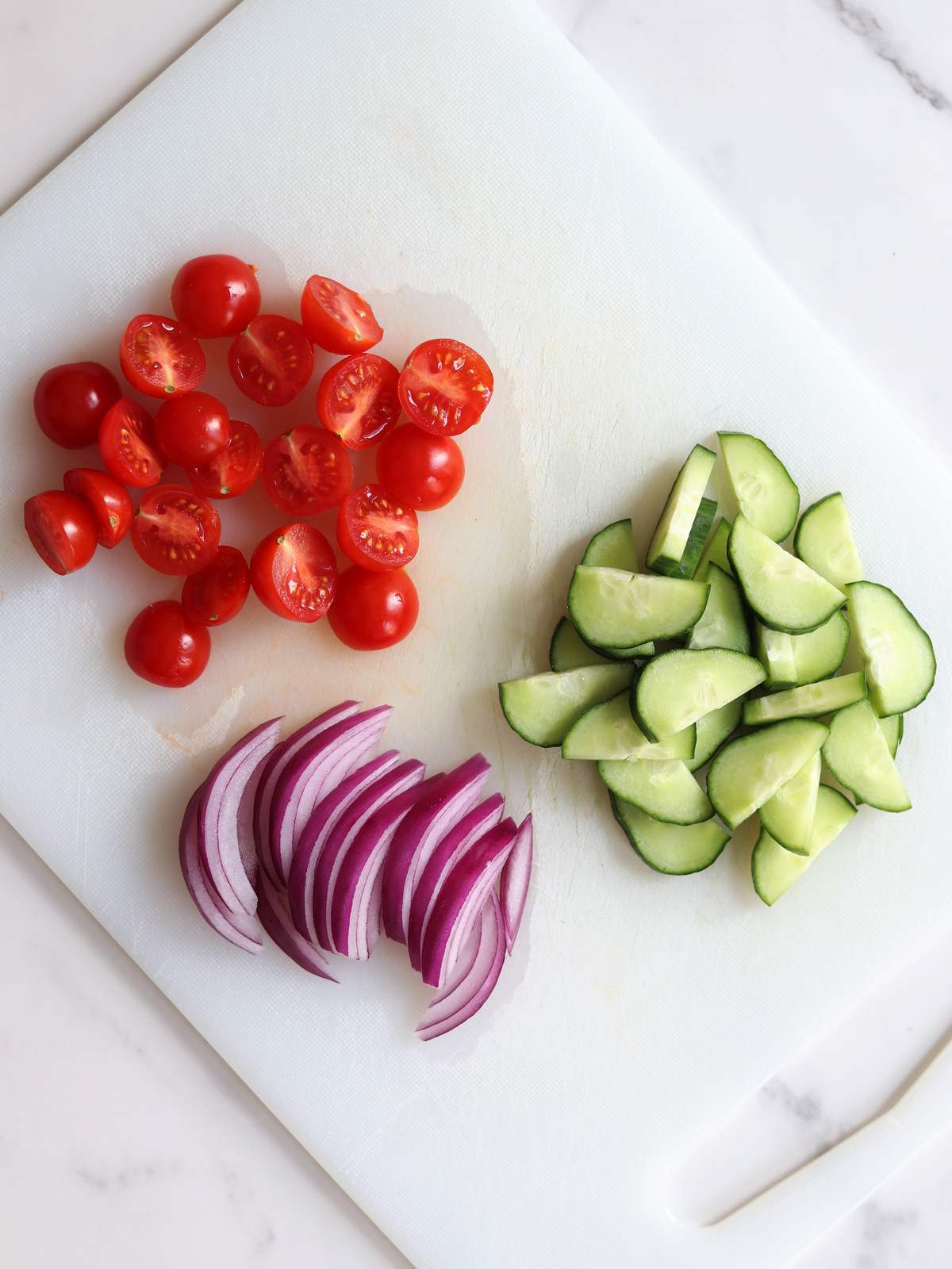 Sliced cherry tomatoes, cucumbers, and red onions are neatly arranged on a white cutting board atop a marble countertop.