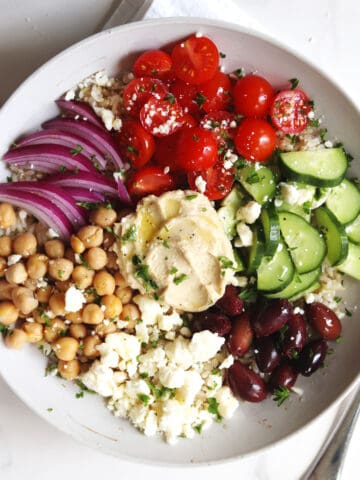 A Mediterranean bowl with chickpeas, feta cheese, Kalamata olives, sliced cucumbers, cherry tomatoes, red onion, and hummus, garnished with fresh parsley.