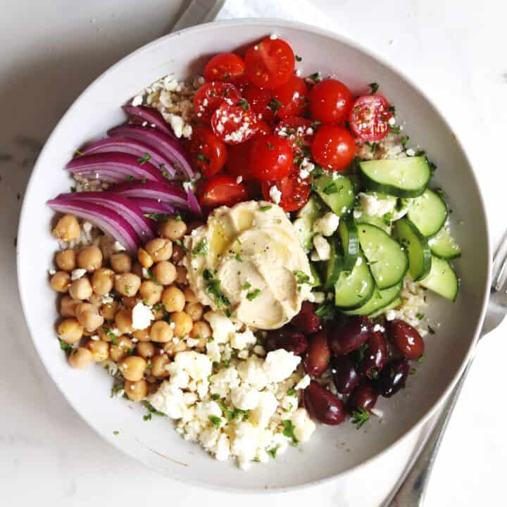 A Mediterranean bowl with chickpeas, feta cheese, Kalamata olives, sliced cucumbers, cherry tomatoes, red onion, and hummus, garnished with fresh parsley.