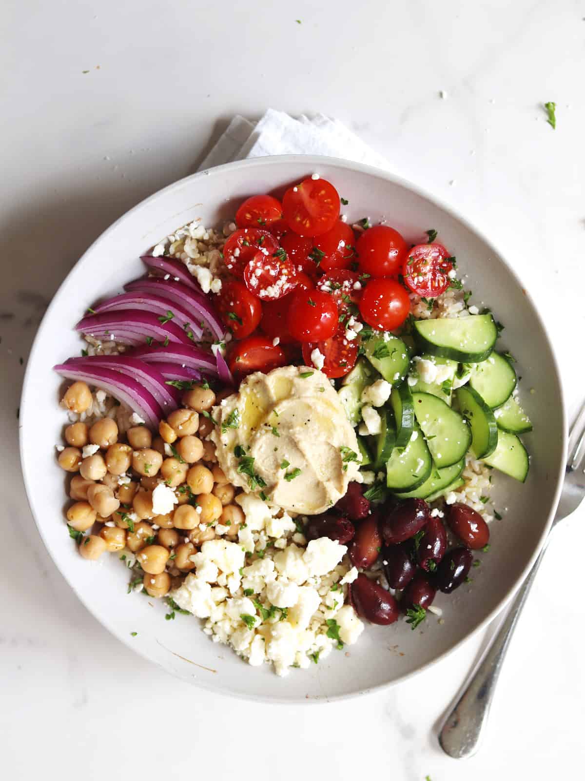 A Mediterranean bowl with chickpeas, feta cheese, Kalamata olives, sliced cucumbers, cherry tomatoes, red onion, and hummus, garnished with fresh parsley.