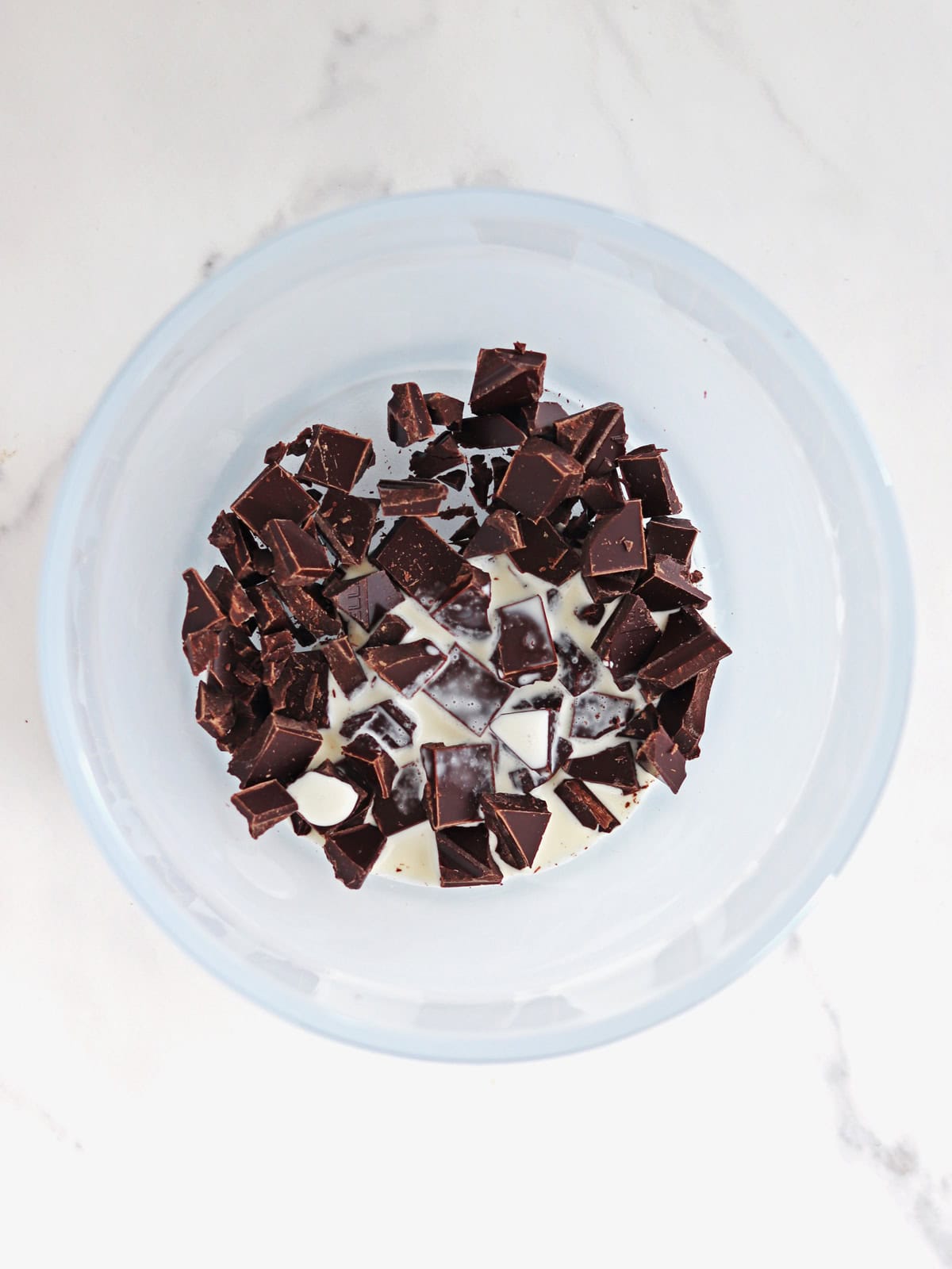 A frosted glass bowl contains chopped semi-sweet chocolate pieces and a small amount of cream, placed on a white marble countertop.
