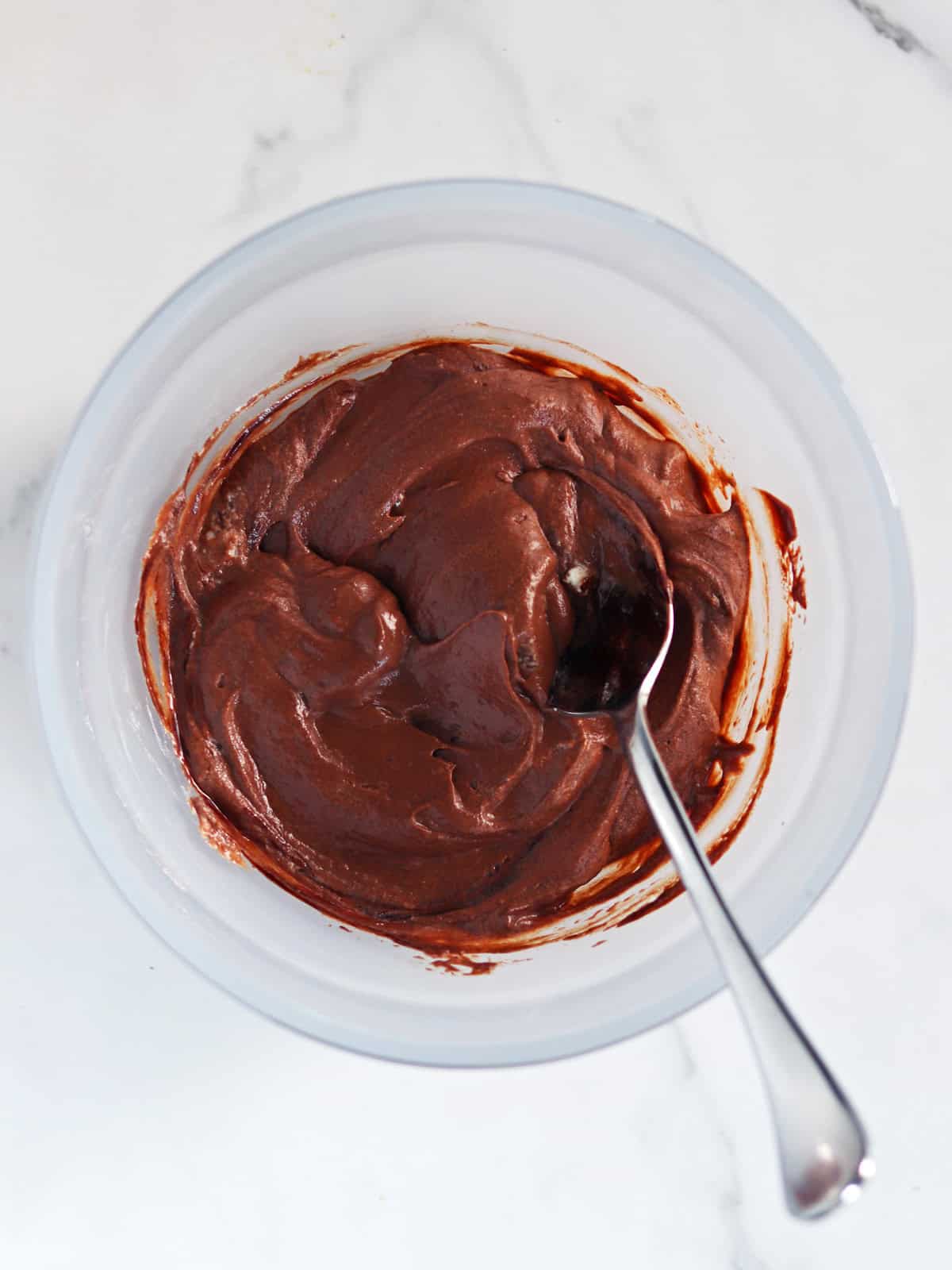 A metal spoon rests in a frosted glass bowl filled with chocolate mousse, sitting on a white marble countertop.