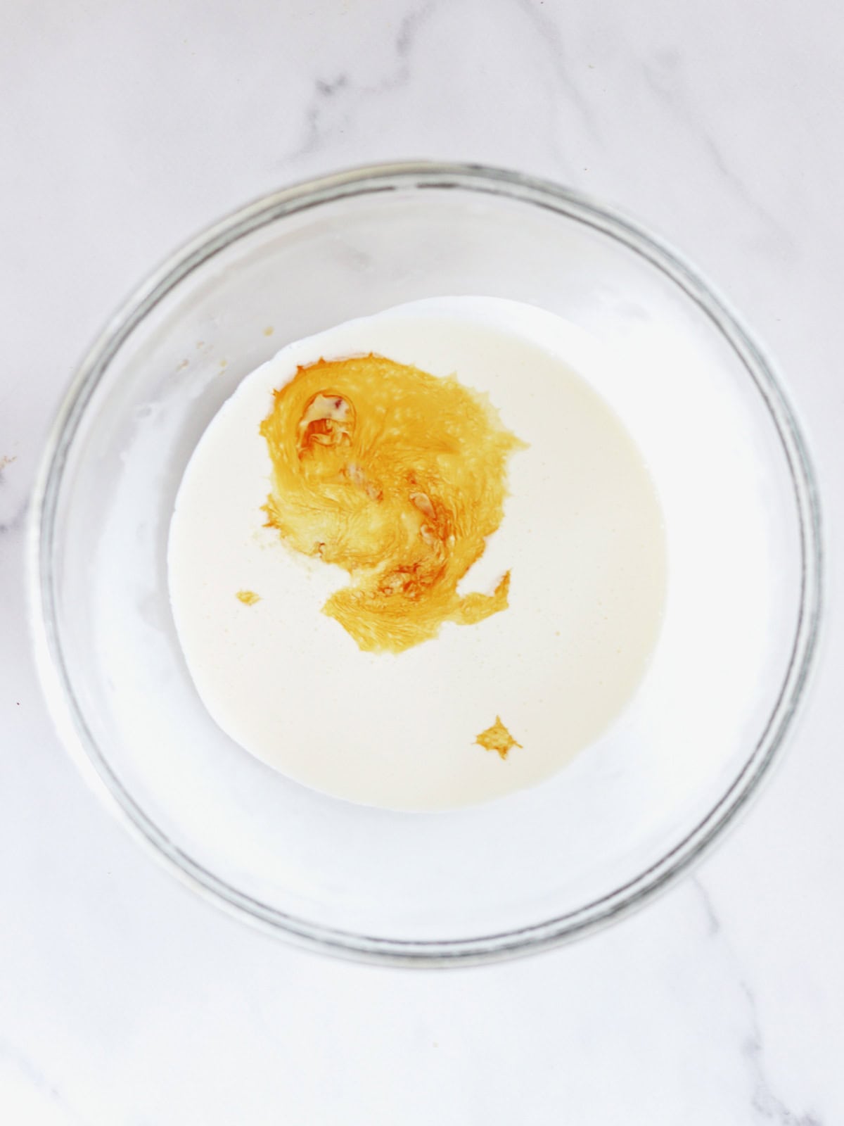 A glass bowl with heavy cream, sugar and vanilla on a white marble countertop, viewed from above.