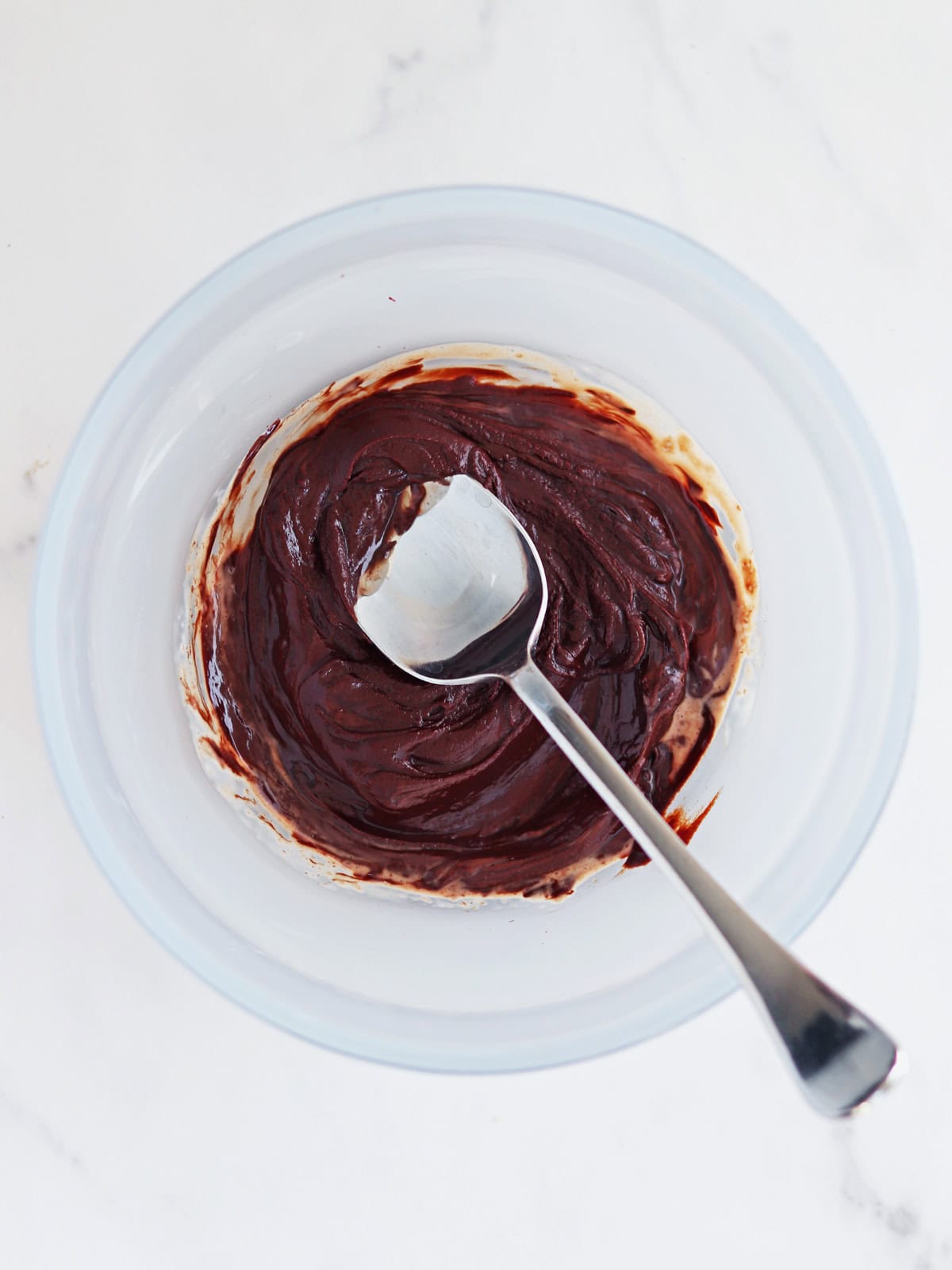 A metal spoon rests in a frosted glass bowl containing melted chocolate and cream, viewed from above on a white marble countertop.