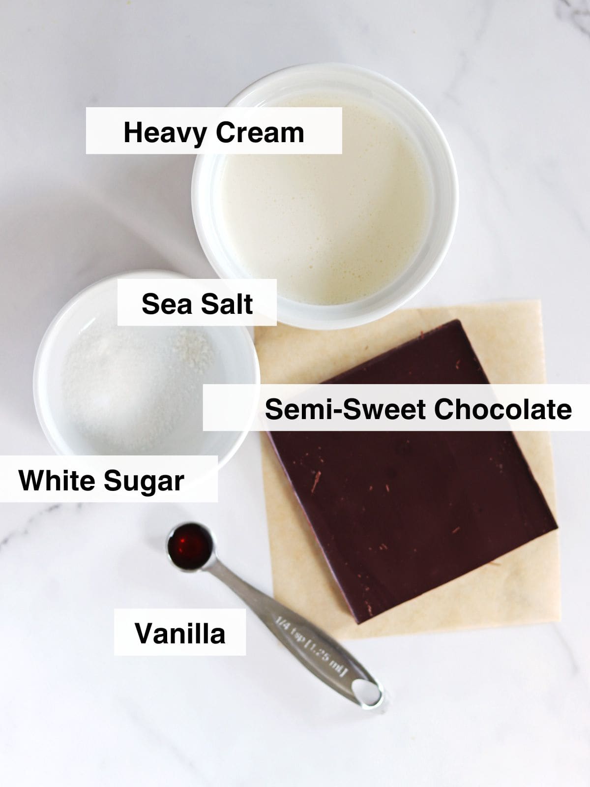Top-down view of labeled ingredients for microwave chocolate mousse: a bowl of heavy cream, a bowl of sea salt and white sugar, a block of semi-sweet chocolate on parchment, and a spoonful of vanilla on a white countertop.