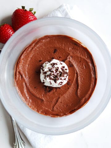 A bowl of chocolate mousse topped with whipped cream and chocolate shavings, with two strawberries, a spoon, and a white napkin beside it on a white countertop.