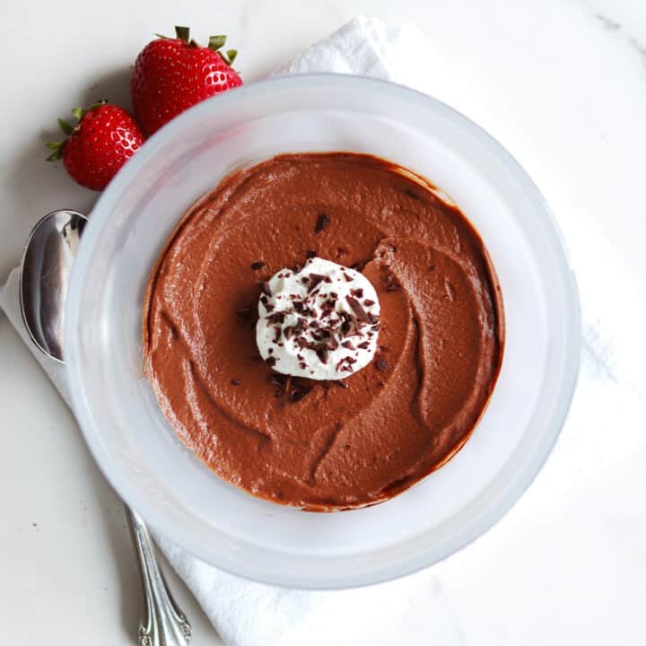 A bowl of chocolate mousse topped with whipped cream and chocolate shavings, with two strawberries, a spoon, and a white napkin beside it on a white countertop.