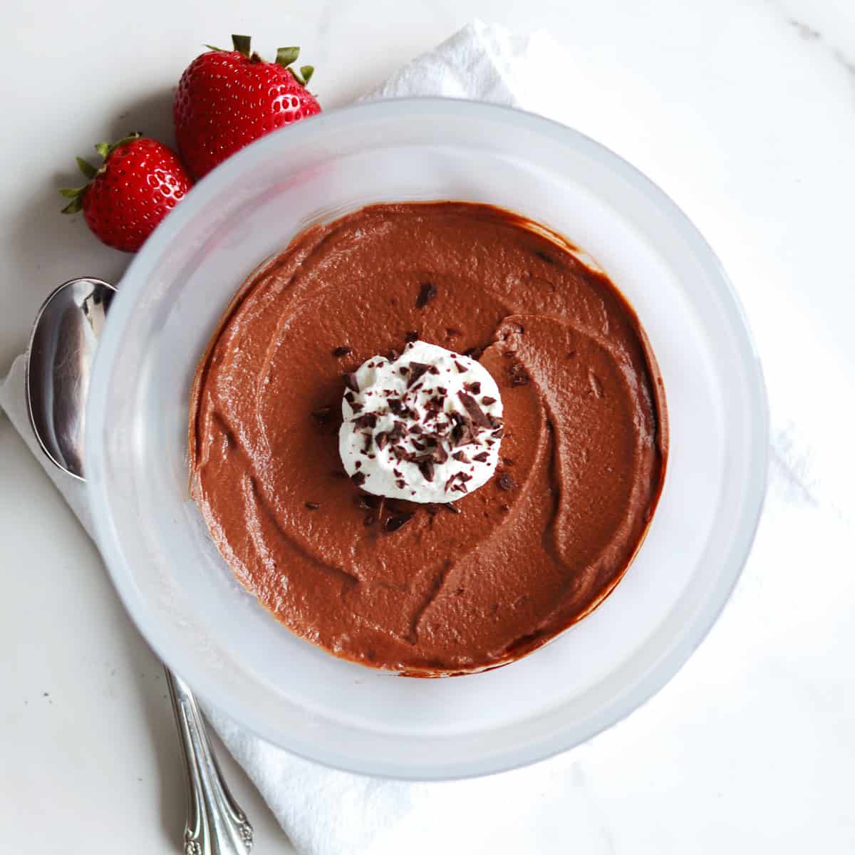 A bowl of chocolate mousse topped with whipped cream and chocolate shavings, with two strawberries, a spoon, and a white napkin beside it on a white countertop.