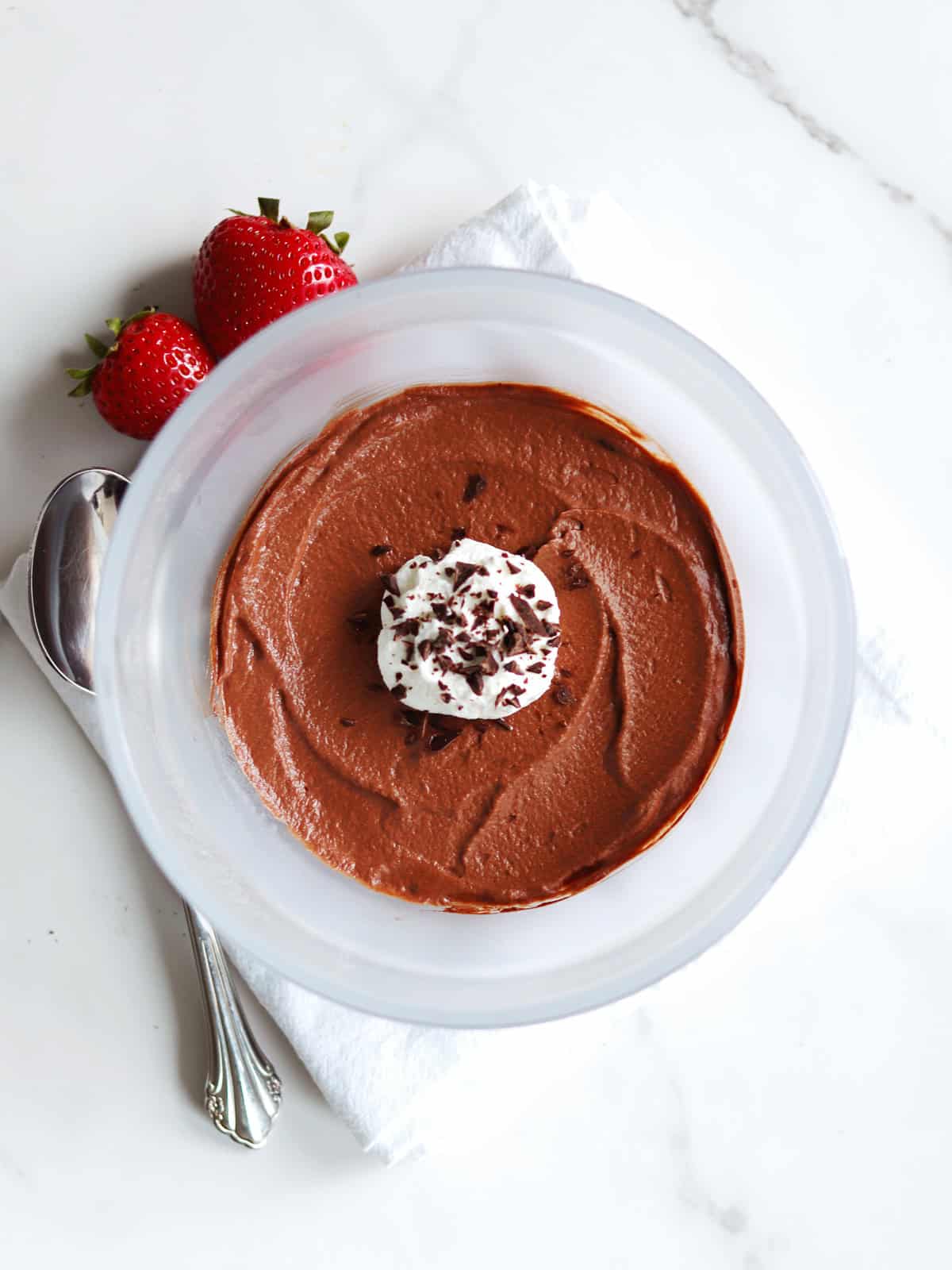 A bowl of chocolate mousse topped with whipped cream and chocolate shavings, placed on a white napkin with a spoon and two strawberries beside it on a marble countertop.