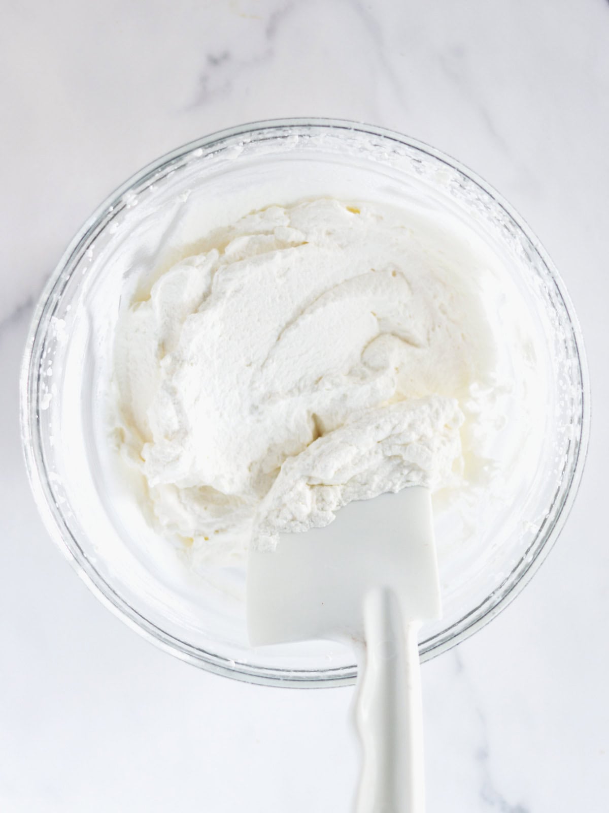 A glass bowl of homemade whipped cream with a white spatula resting inside, on a white marble countertop.