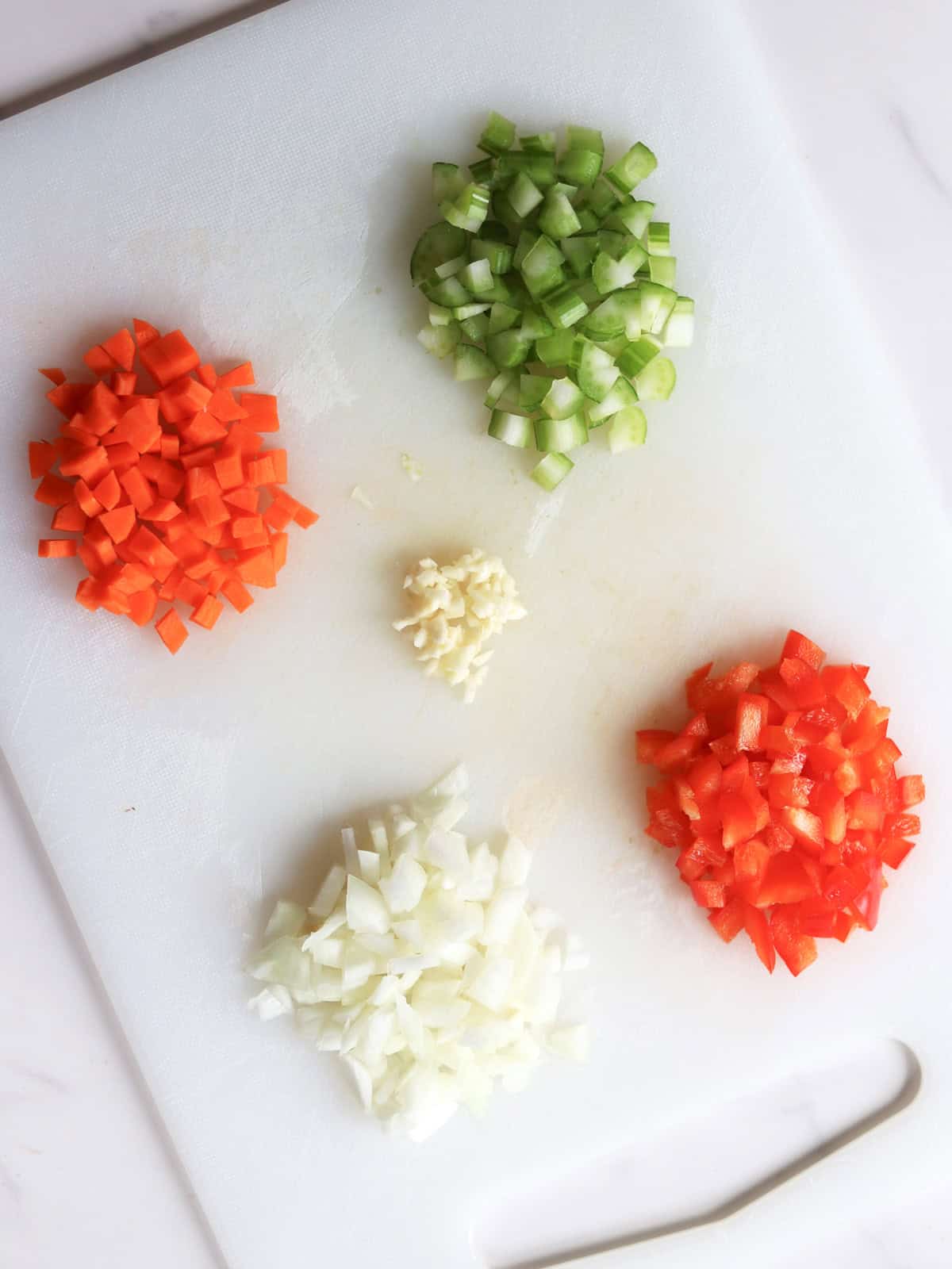 Chopped vegetables on a white cutting board: carrots, celery, onion, red bell pepper, and minced garlic are arranged in separate piles.